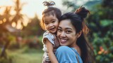 Fototapeta  - Asian mom and her cute little daughter, woman carrying the female child or kid in her hands, both of them are smiling and looking at the camera. Walking in nature, happy family, motherhood concept
