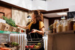 Woman in low carbon footprint zero waste store interested in purchasing food with high nutritional value. Client fills shopping basket with pesticides free produce in local neighborhood shop