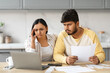 © Prostock-studio - Overwhelmed couple sitting together at kitchen, using laptop, holding papers