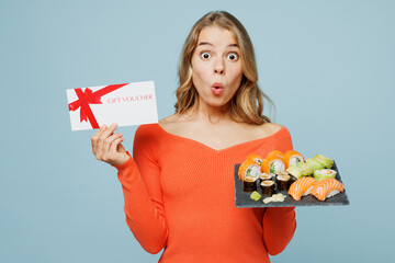 Young surprised woman wearing orange casual clothes hold store gift coupon voucher card eat raw fresh sushi roll served on black plate Japanese food isolated on plain blue background studio portrait.