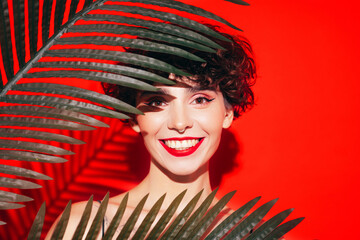 Fashion beauty portrait of young smiling brunette woman with evening stylish  makeup and perfect clean skin. Sexy model with curly short hair posing in studio. With red bright lips. Tropical palm leaf