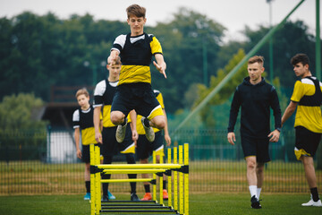 Wall Mural - Football camp for youth. Teenage boys in training with a coach at grass pitch. Sports team preparing for the season. Young players jumping over hurdles for strength and coordination training