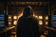 Back view of unrecognizable man in hoodie standing near desk and reading stolen data from computer, monitors in dark room before massive cyber attack on servers.