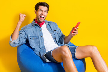 Photo of ecstatic funky guy wear jeans jacket shorts sit on pouf hold smartphone win betting isolated on bright yellow color background