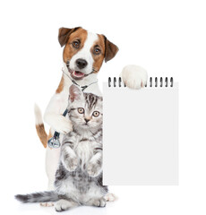 Sticker - Smart jack russell terrier with stethoscope on his neck hugs tiny kitten and shows empty notebook. isolated on white background