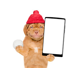 Poster - Happy puppy wearing a warm hat with pompon holds snowball and holds smartphone with white blank screen in it paw. isolated on white background