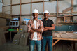 © Prins Productions - Male African colleagues with hardhats stand smiling in carpentry factory with arms folded
