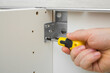 Young adult man hand using manual screwdriver and screwing screw in white board and metal angle corner for base cabinet frame fastening on wall rail. Assembling new kitchen wooden furniture. Closeup.