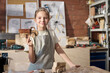 Waist up portrait of smiling young girl posing in carpentry workshop and holding handmade wooden toy