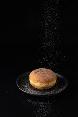 Wall Mural - Single fresh baked donut sprinkled with falling sugar powder on dark retro plate on black background. Berliner donut.