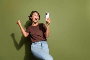 Wall Mural - Photo portrait of attractive young woman raise fist win hold device excited wear trendy brown clothes isolated on khaki color background