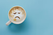 Coffee cup with sad face drawn on coffee milk foam. Top view to mug with coffee on blue background. Blue monday, hard morning, difficult day, negative emotions, loneliness, loss, problem, difficulties