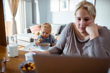 Busy Mother Working On Laptop While Feeding Toddler At Home