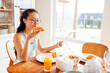 Young attractive Asian woman in glasses and casual clothes is sitting at table and eating sandwich. Charming girl is having breakfast in kitchen at home.