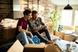 Young male gay couple looking at smartphone with moving boxes in new home