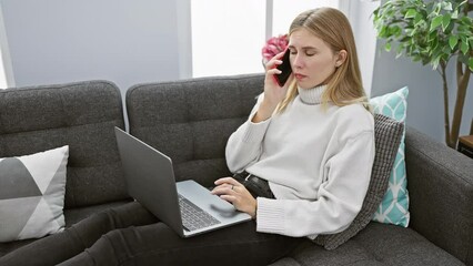 Poster - Blonde woman converses on phone while using laptop on gray couch indoors, displaying multi-tasking and modern lifestyle.