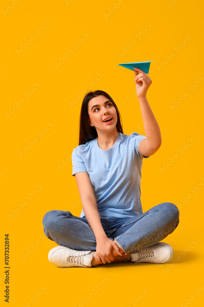 Young woman with paper plane sitting on yellow background