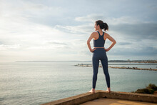 Rear view of a sporty woman facing ocean with hands on hips, relaxing after exercise.