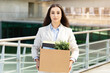 © Prostock-studio - A composed professional woman carrying a cardboard box filled with office supplies and a plant