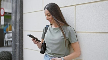 Wall Mural - Cheerful, young hispanic woman typing an enchanting message on her cell phone, leaning on an urban street wall, expressing the joy of digital communication.