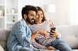 © Prostock-studio - Happy indian couple sitting on sofa at home, using smartphone