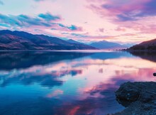 Serene Lake At Sunset With Vibrant Pink And Purple Sky Reflecting On Calm Water, Surrounded By Mountains.