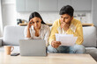 © Prostock-studio - Upset indian couple checking papers and using laptop at home