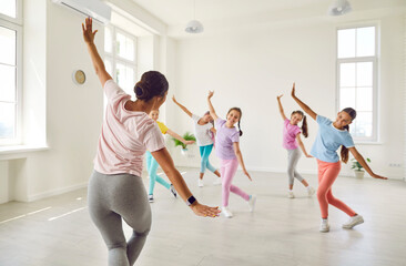 Wall Mural - Happy children having dance class with woman teacher. Kids learning some ballet like moves. Group of little girl dancers rehearsing new choreo in spacious light white room at modern gym or dance hall