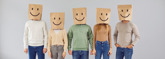 Portrait five casual male and female people with paper shopping package bags over heads with happy smiling emoticon mouth eyes facial expression drawn on them standing on gray color studio background