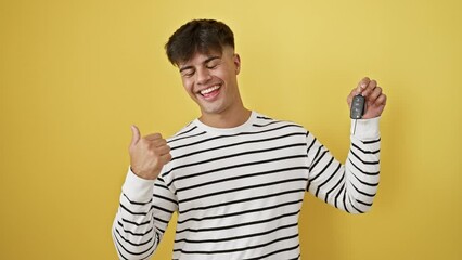 Canvas Print - Joyful young hispanic man holding car key, pointing to side with thumbs up, smiling wide with an open mouth, backed by a sunny yellow, isolated background.