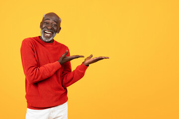 Wall Mural - Cheerful elderly Black man in red sweater showcasing with hands on an empty space