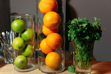 Fresh Citrus Fruits and Mint in Glass Containers. Bright oranges and limes stacked in clear glass vases with a bouquet of fresh mint leaves on a wooden counter, ready for cocktail making.