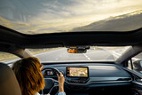 Fototapeta Sypialnia - Woman drives car with panoramic rooftop on sunset. Interior view from a backside. Modern cars and travel concept