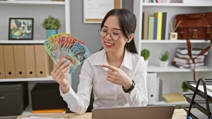 Sticker - Joyful young chinese woman excitedly points out australian dollars in office, smiling with hopeful confidence