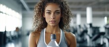 Tanned Young Student Girl In Sportswear Holds Bottle Looks At Camera With Wrapped Towel Around Neck At Fitness Club.