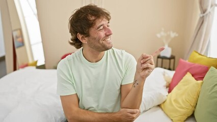 Wall Mural - Cheerful young caucasian man in pyjama confidently pointing to the side, big smile lighting up his face, looking straight at the camera from his cozy bedroom