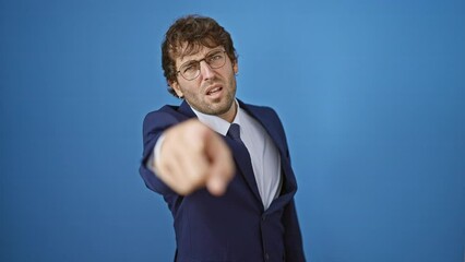 Canvas Print - Displeased young man in business suit, furious anger blazing, pointing a blame finger at you through the camera, isolated on blue background.