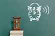 A vintage hourglass standing on a book against the backdrop of a chalkboard with an alarm clock drawn in chalk. There is copy space on the board for inserting text or drawing