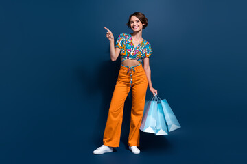 Poster - Full size photo of attractive young woman shopping bags point empty space wear trendy flared pants isolated on dark blue color background