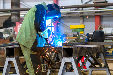 A welder wearing a protective mask for metal welding and protective gloves performs welding work at a metal structures plant. Welding a steel industrial beam. Sparks, hot flame from welding.