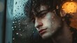 A young man is staring out of a window with raindrops on it, looking contemplative or somber.