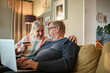 © Marko Geber - Happy elderly couple sitting on couch using credit card on laptop
