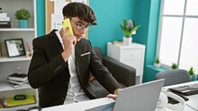 Dedicated young hispanic teenager worker talks business over smartphone, working at office laptop, defining success in his professional indoor room.