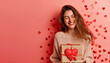 Happy young woman celebrating Valentine`s Day, adult girl holds gift box on pink studio background, small heart shapes design. Concept of love, party, fun, smiling people