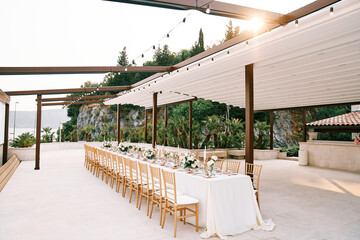 Wall Mural - Long set table with a white tablecloth and bouquets of flowers stands surrounded by chairs on a garden terrace under a canopy