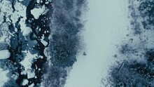 Aerial top down view of a solo male person walk along the snowy frozen river, surrounded by pine trees. Cold winter forest. National park ranger in a fur coat in a cold winter scenery.