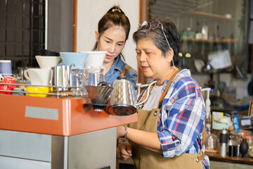 Wall Mural - Young Asian barista female smiling happy working with retired mother bonding relationship, cafe shop small business entrepreneur woman teaching 60s senior elderly pensioner worker making hot coffee