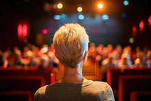 A Woman Watches A Live Performance Or Event, Sitting In A Theater Audience With Stage Lights Visible Ahead.