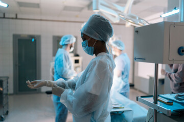 Wall Mural - African-American female nurse in mask putting on disposable gloves as hygiene before surgery in hospital on background of male surgeons. Concept of preparing for surgical operation.