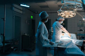 Wall Mural - Remote shot of male surgeon and African-American female nurse performing operation under bright electric lamp in modern operation theatre. Doctors wearing sterile gloves, surgical uniforms and masks.